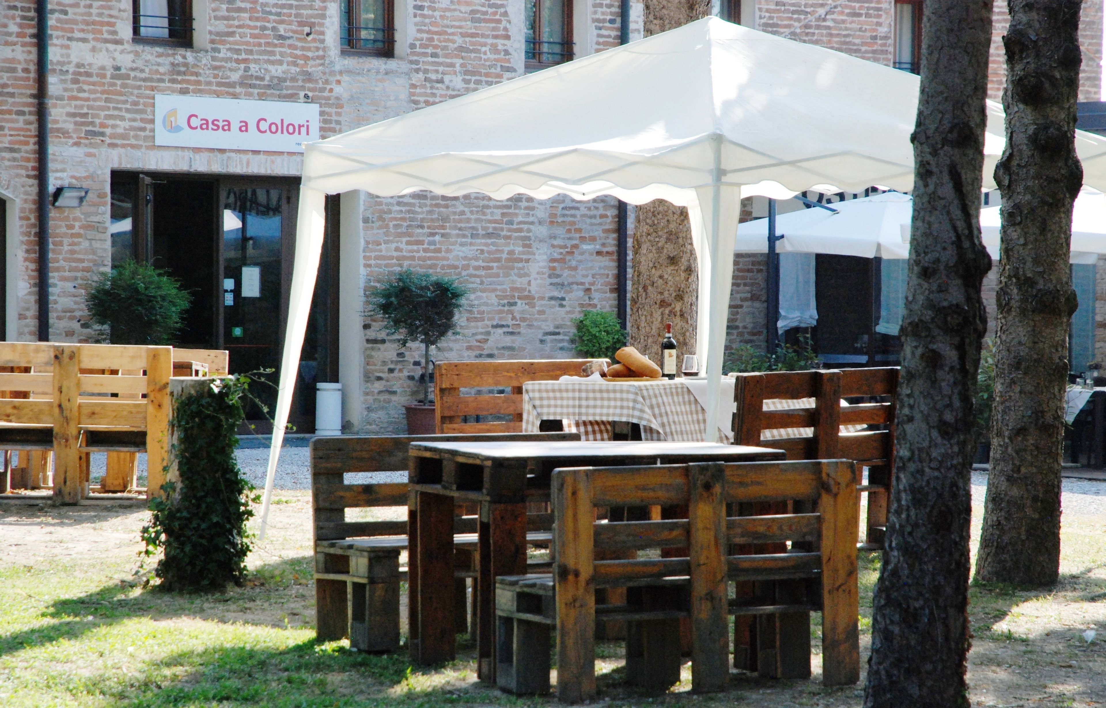 Un área de estar al aire libre con mobiliario de madera bajo un toldo blanco, un edificio de ladrillo al fondo y un letrero que dice "Casa a Colori".