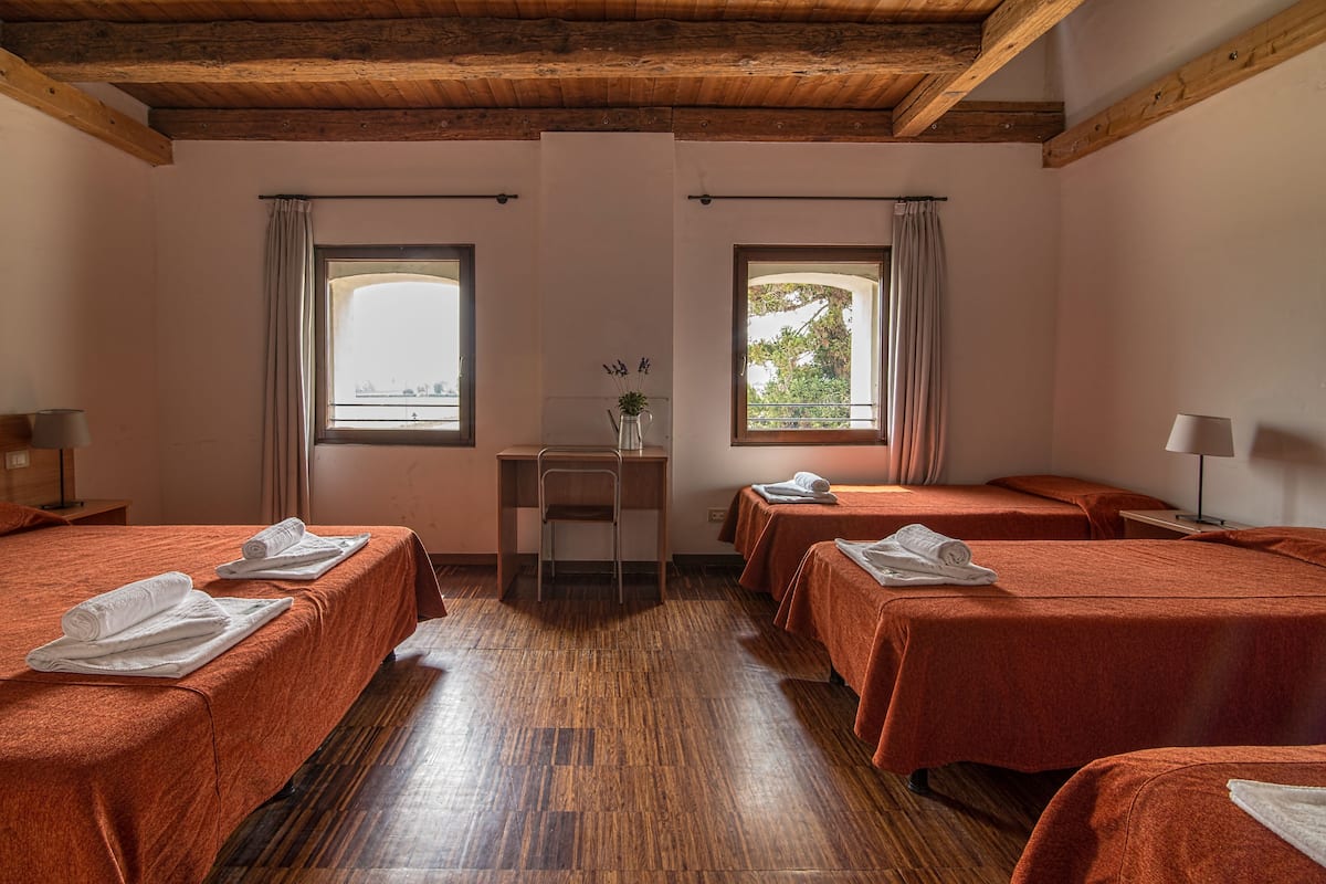 A hotel room with four beds, wooden flooring, a desk with a chair, and two windows with curtains.