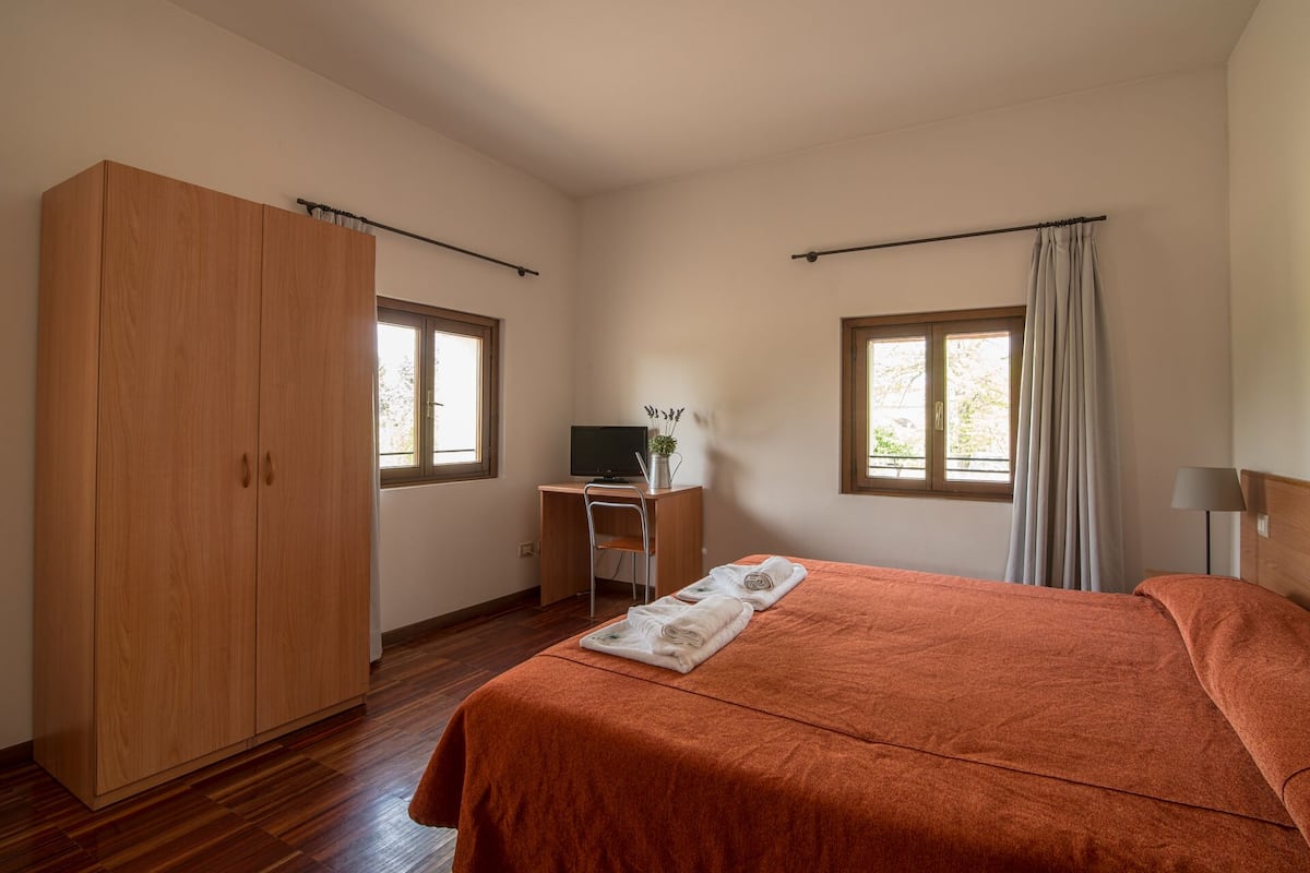 A bedroom with a bed, a desk with a computer, a wardrobe, and two windows with curtains.