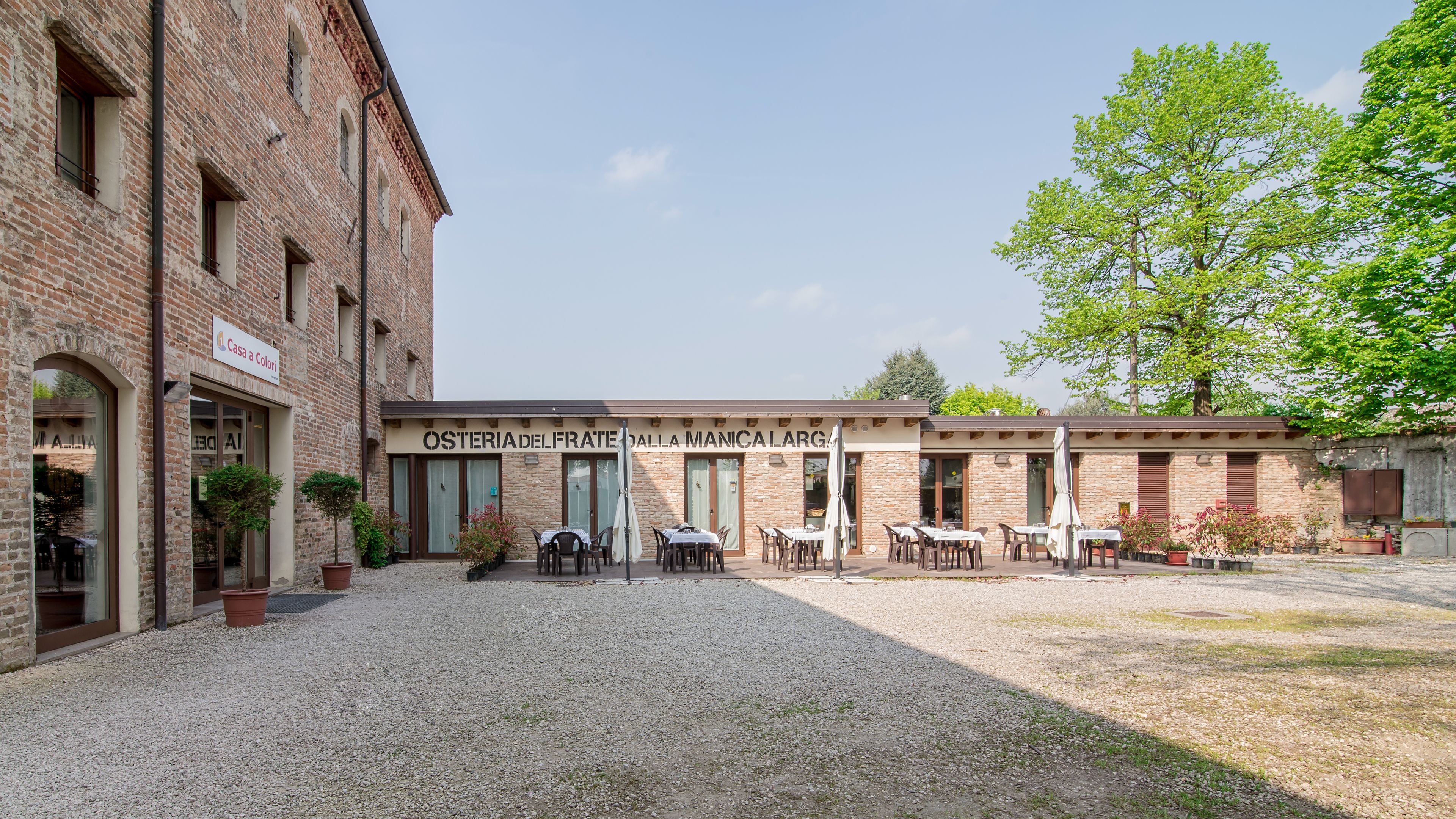 A courtyard with outdoor seating, a brick building, and a sign for 'Osteria del Frate Dalla Manica Larga'.