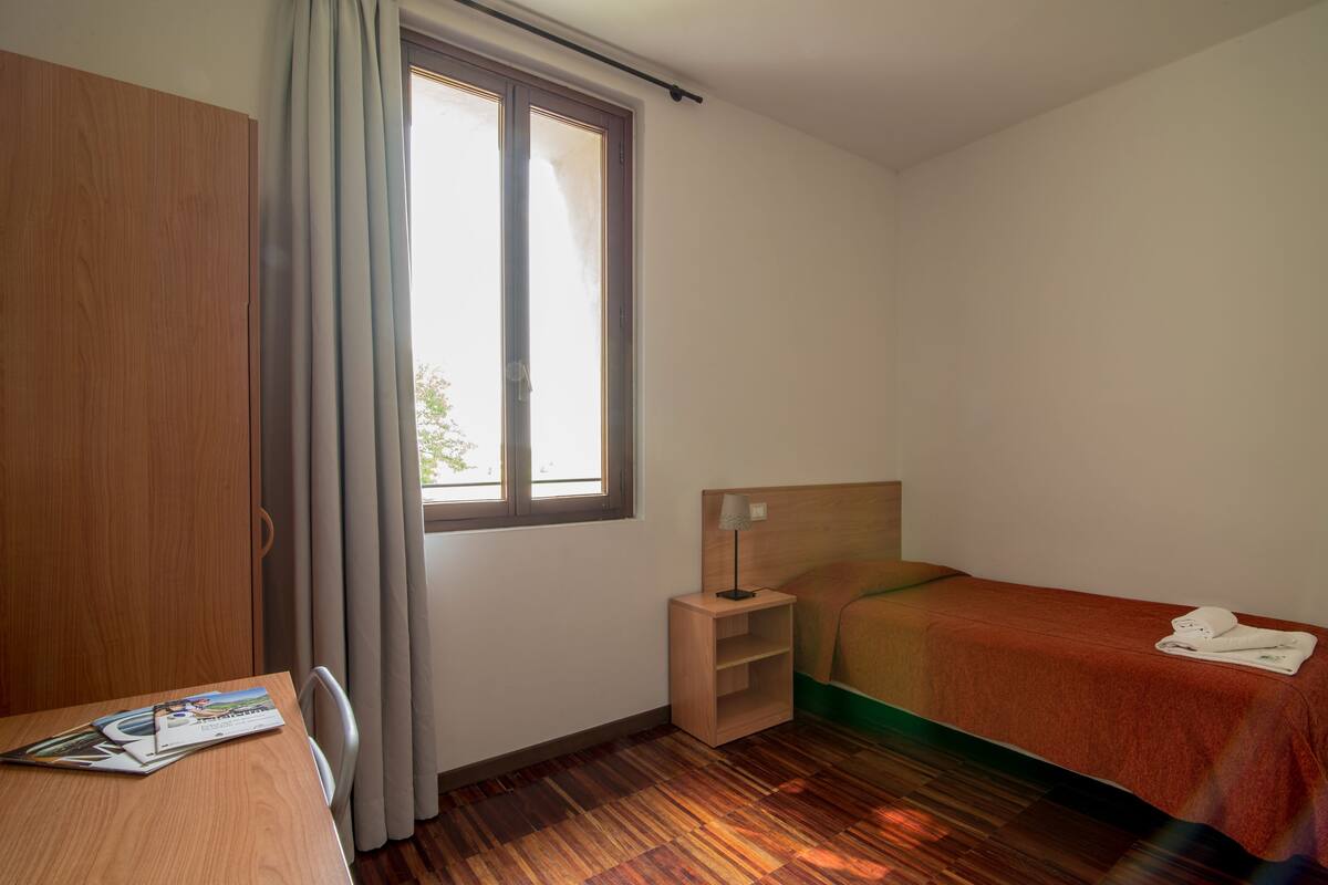 A hotel room with a wooden wardrobe, a small bedside table with a lamp, a single bed with a red blanket, and a window with curtains.