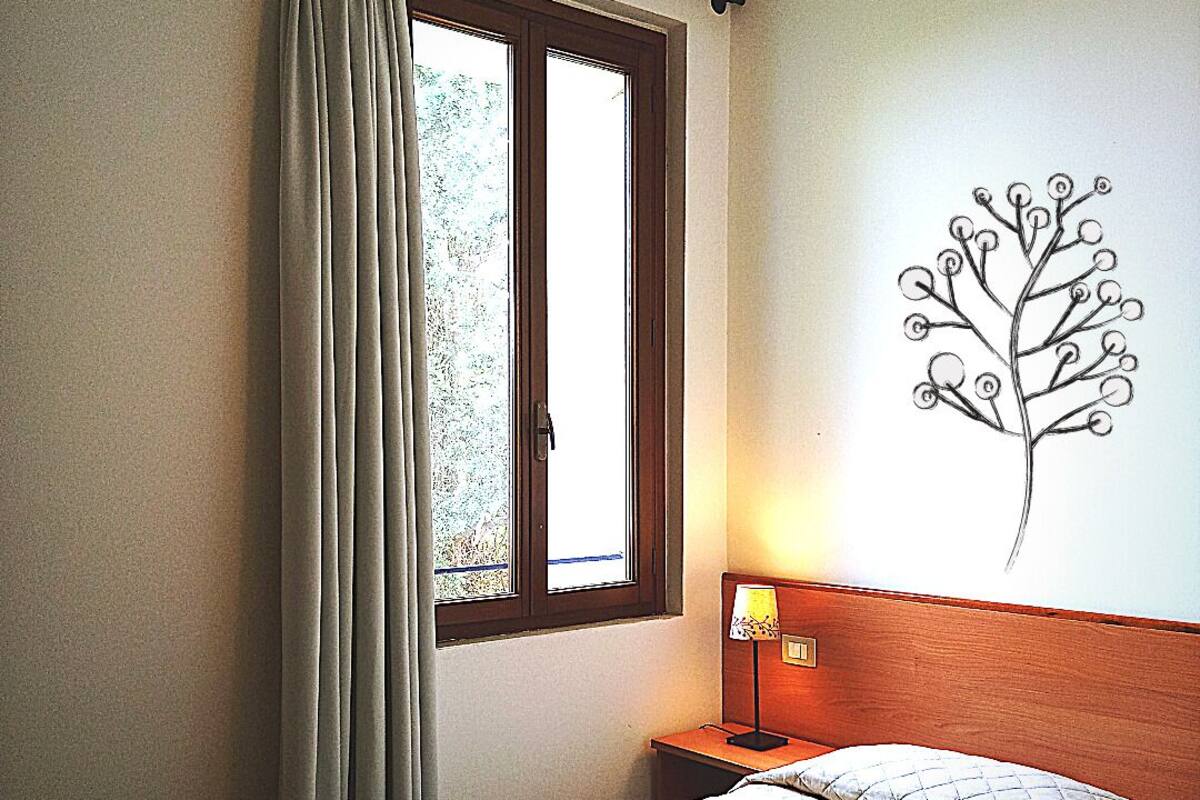 A hotel room with a bed, a window with curtains, a nightstand with a lamp, and a wall art piece.