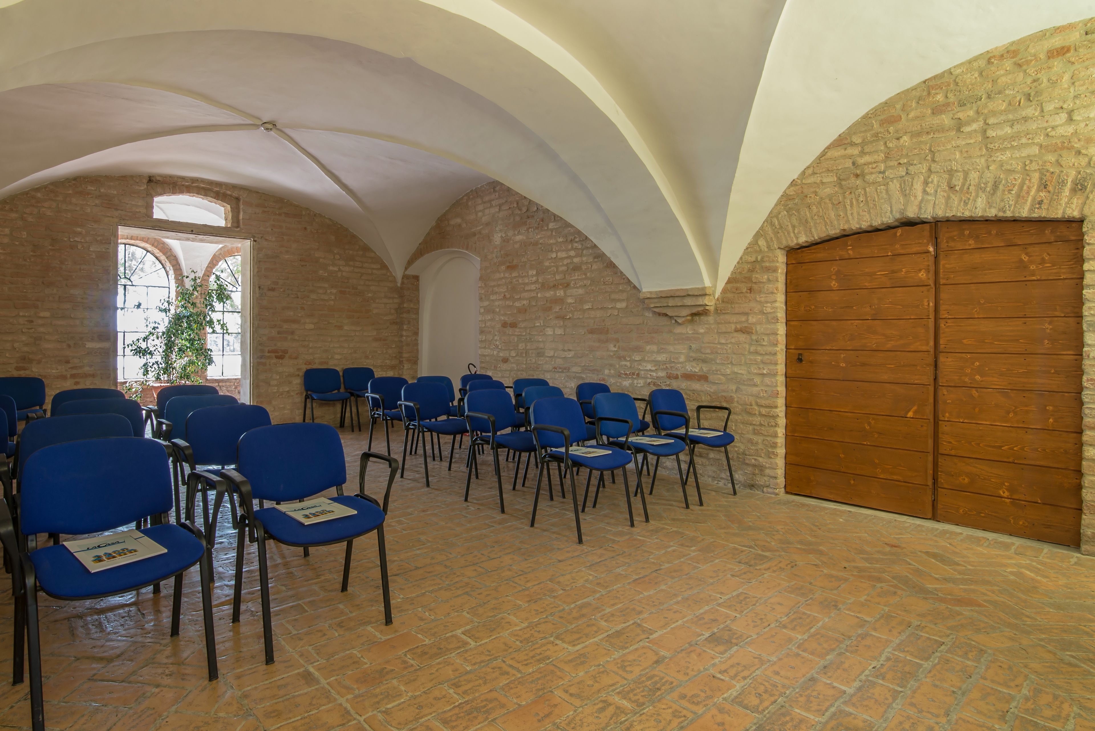 Una stanza con pareti in mattoni, soffitti a volta e un'ampia porta in legno. File di sedie blu disposte per i posti a sedere.