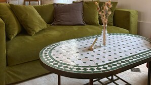 A cozy living room with a green sofa, a round table with a mosaic top, and a kitchen in the background. - Hostellerie Du Lys - Hôtel & Restaurant (Lamorlaye)