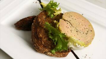 Un plato rectangular blanco con un trozo de pan marrón oscuro cubierto de lechuga verde y una porción de albóndigas rosadas, acompañado de salsa oscura y un pequeño bocado frito.