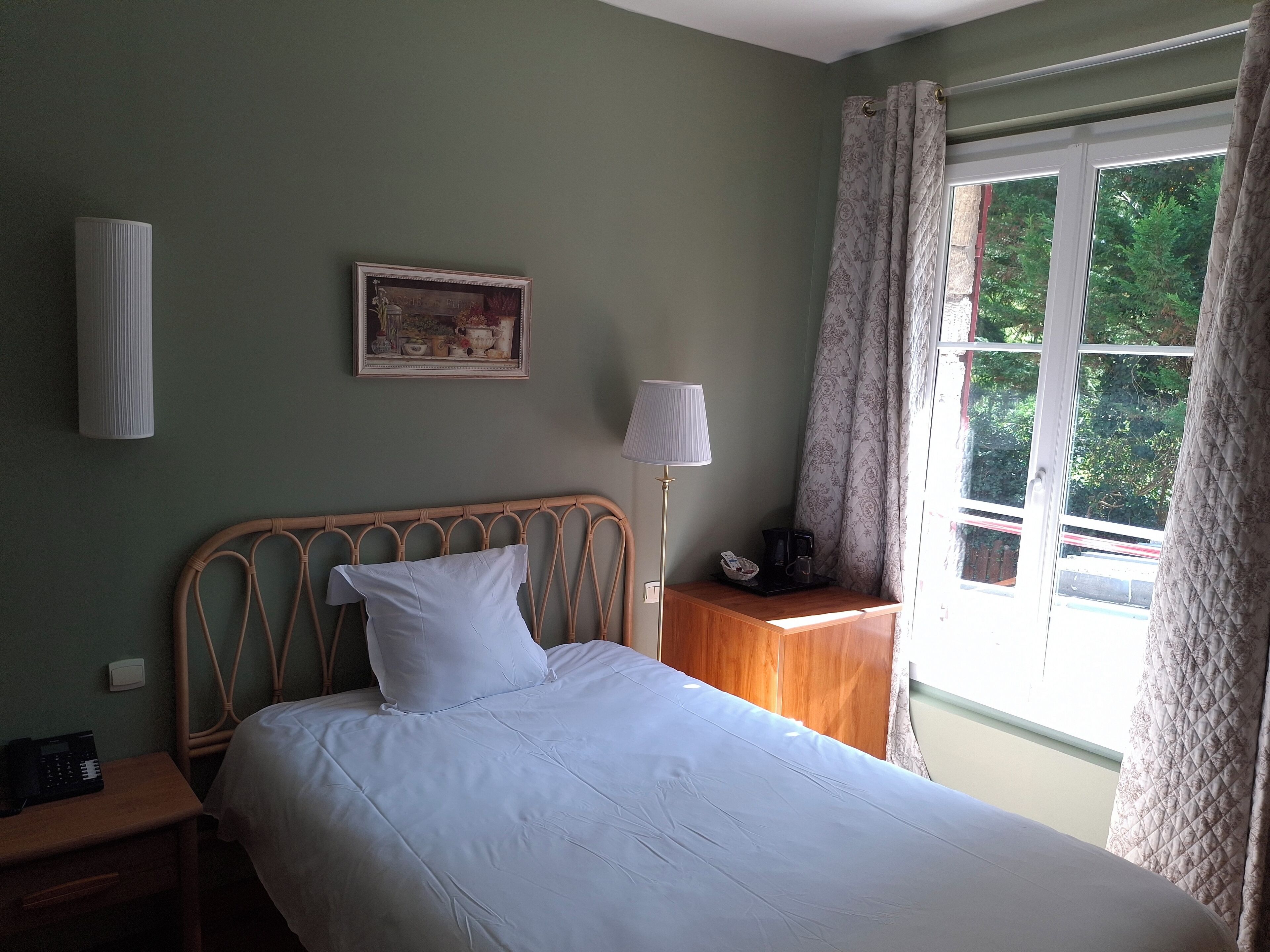 Een netjes opgemaakt bed met witte lakens, een bedlampje, een klein houten nachtkastje met een telefoon, en een raam met gordijnen.