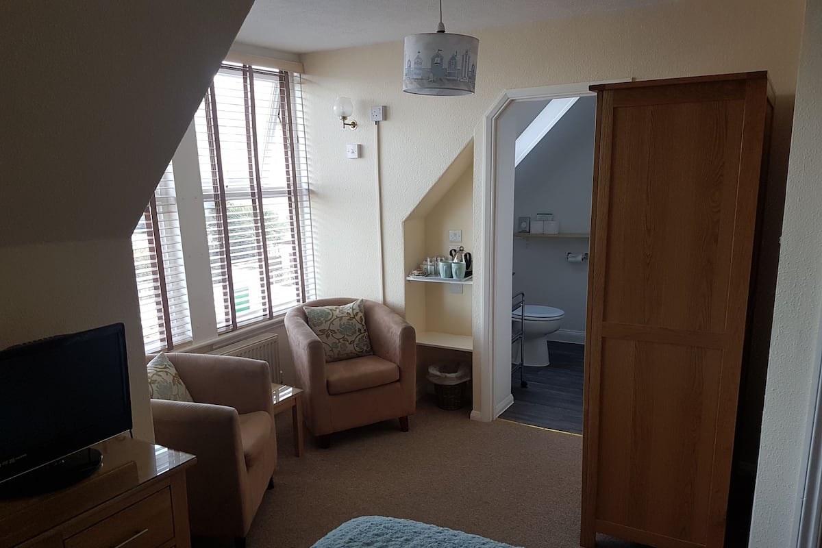 A bedroom with a TV, a sofa, a chair, a bed, a wardrobe, and a bathroom visible through an open door.