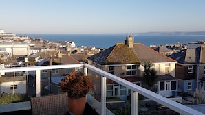 A balcony with wicker furniture and a potted plant, overlooking a coastal town with houses and a view of the sea. - Trelinda Hotel (Newquay)