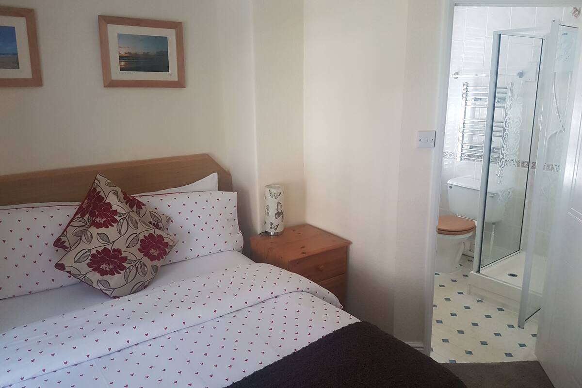 A bedroom with a bed, a nightstand, a bedside table, a mirror, and a bathroom with a shower and toilet.