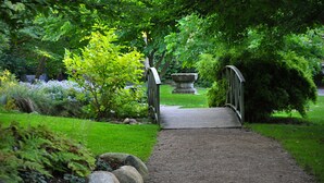 A gravel pathway leading to a wooden bridge over a small stream, surrounded by lush greenery and a stone planter. - Hotell Lindgården (Visby)
