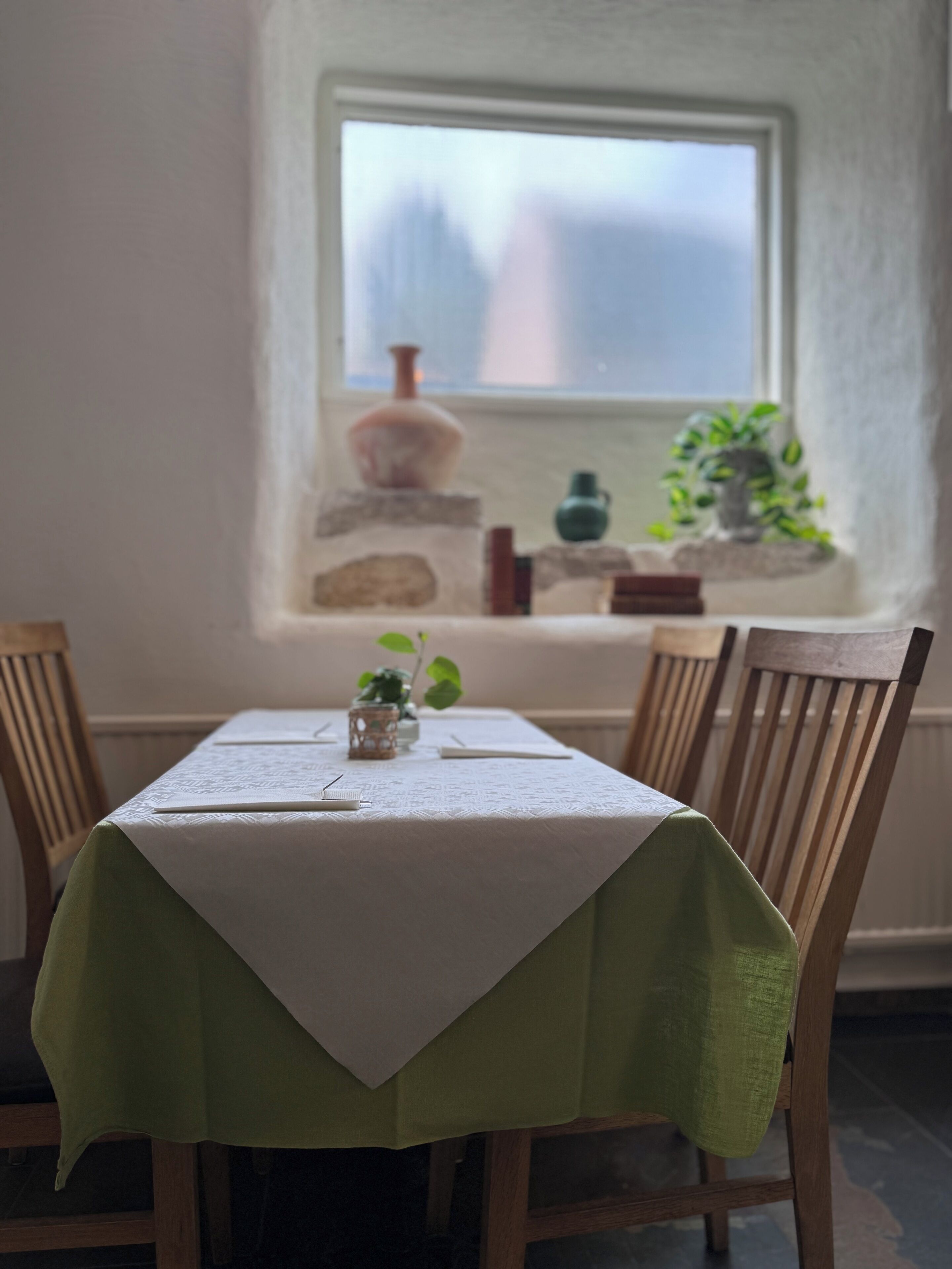 Una mesa de comedor con mantel blanco, camino de mesa verde y cuatro sillas de madera. Un jarrón y una pequeña planta están sobre el alféizar de una ventana.