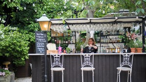 A bar with a sign, a woman behind the bar, and three metal chairs in front. - Hotell Lindgården (Visby)