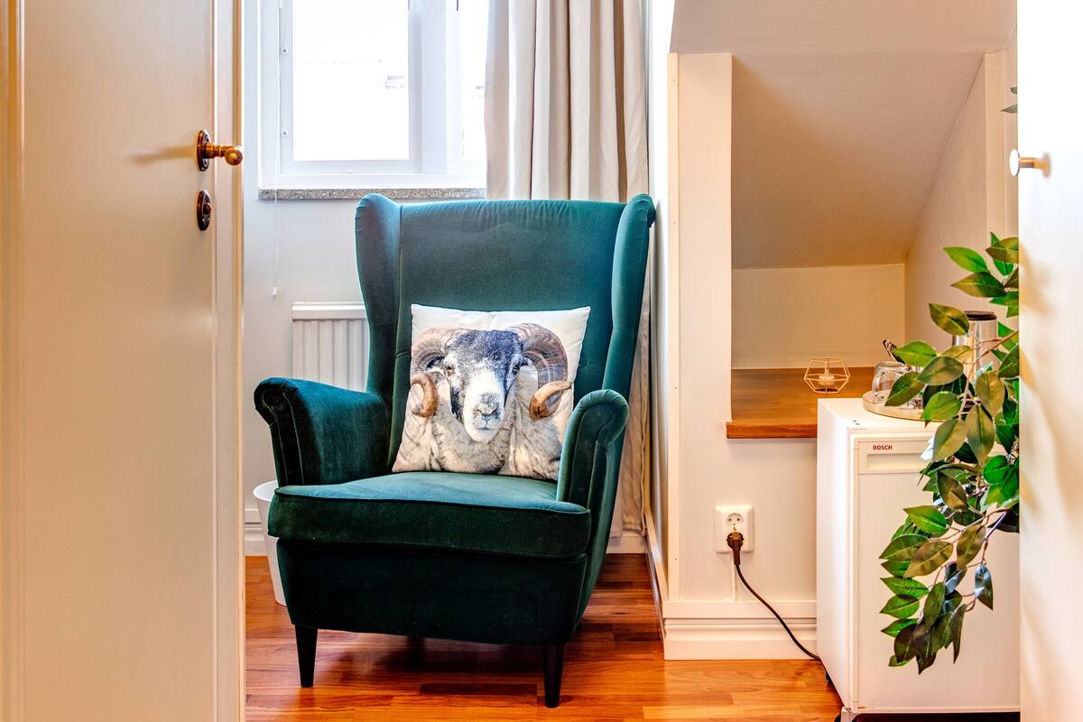 Una habitación con piso de madera, un sillón verde con un cojín decorativo, un refrigerador blanco Bosch y una ventana con cortinas.