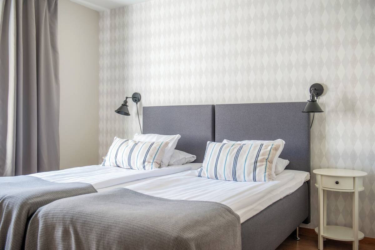 Una cama doble con ropa de cama gris y almohadas a rayas, una mesita de noche con lámpara y un papel tapiz estampado.