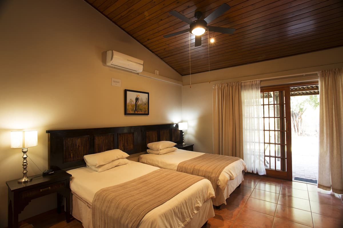 A hotel room with two beds, a ceiling fan, an air conditioning unit, a nightstand with a lamp, and a window with curtains.