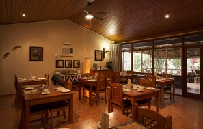 Un comedor con mesas y sillas de madera, un ventilador de techo y vistas amplias a través de grandes ventanas.