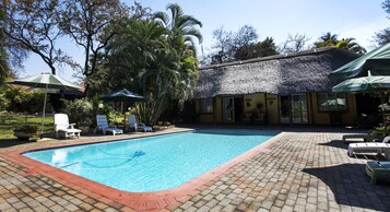 Una piscina con sillas de descanso y sombrillas, rodeada de palmeras y un edificio con techo de paja.
