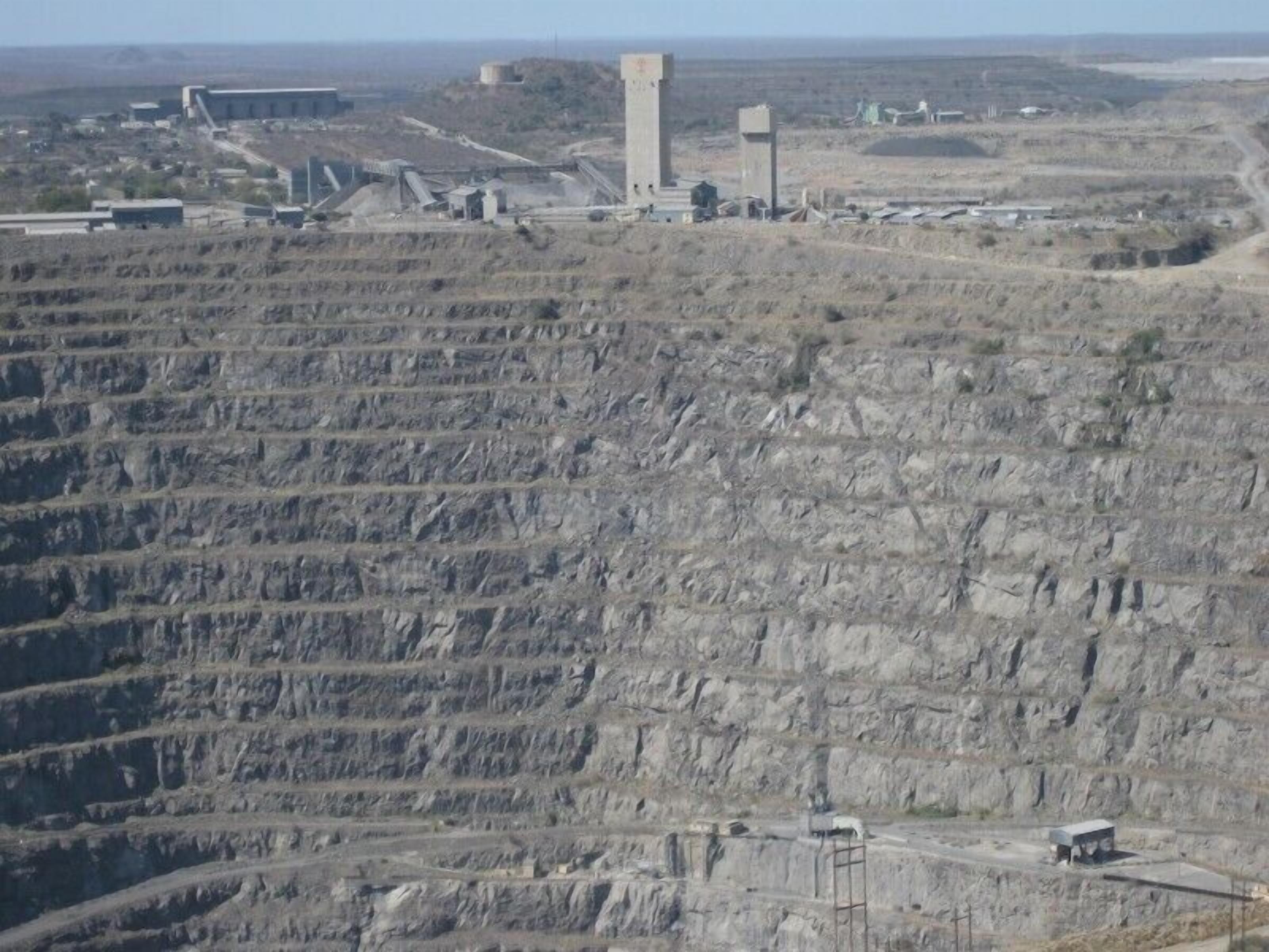 A large open-pit mine with terraced excavation levels and industrial buildings in the background.