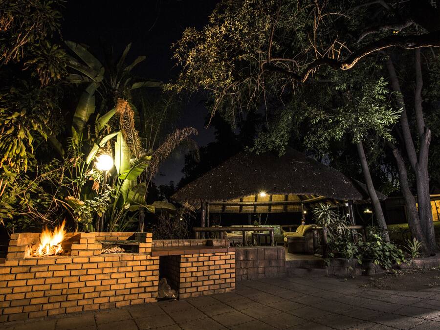 A nighttime outdoor area with a brick fire pit, a thatched-roof structure, and surrounding greenery.