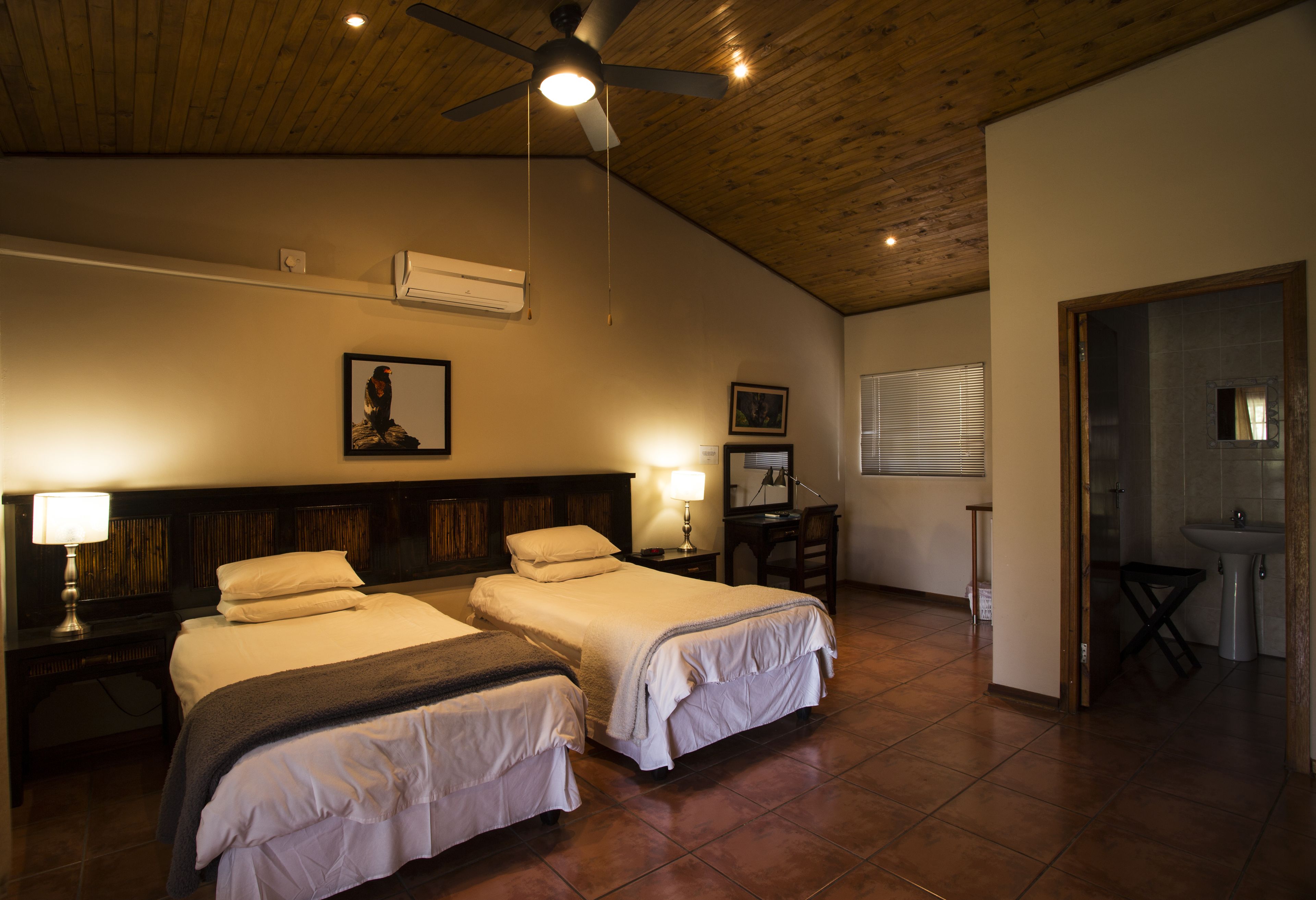 A hotel room with two beds, a ceiling fan, a wooden ceiling, and a bathroom visible through an open door.