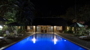 Ein Schwimmbecken mit Liegestühlen und Sonnenschirmen, umgeben von tropischen Pflanzen und einem Gebäude mit einer überdachten Terrasse.