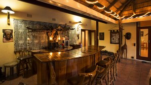 Eine Bar mit Holztresen, Hockstühlen und einer Wand mit Muster. Es gibt Wandleuchten und eine Decke mit einer Strohstruktur.