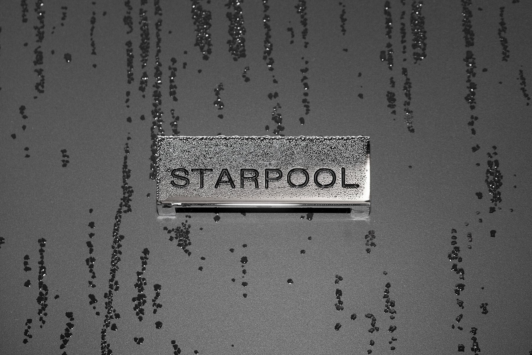 Un objeto metálico con la inscripción “STARPOOL” sobre una superficie texturizada y salpicaduras dispersas.