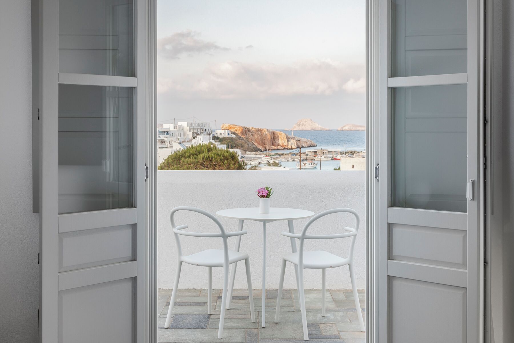 An open balcony with white chairs and a small table, overlooking a coastal town with buildings and cliffs.