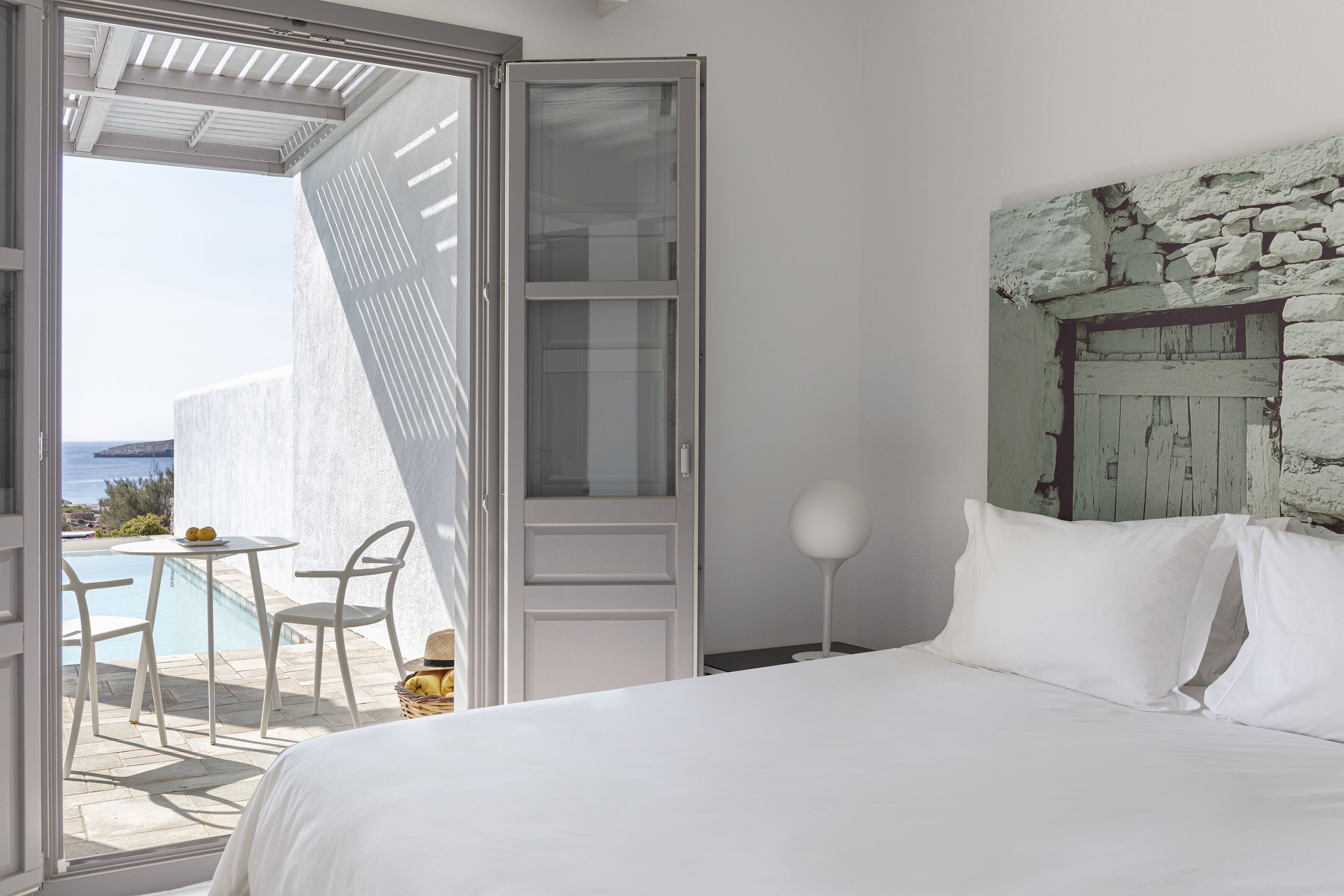 A bedroom with a bed, a white wall, a stone headboard, and a view of the sea through an open door.