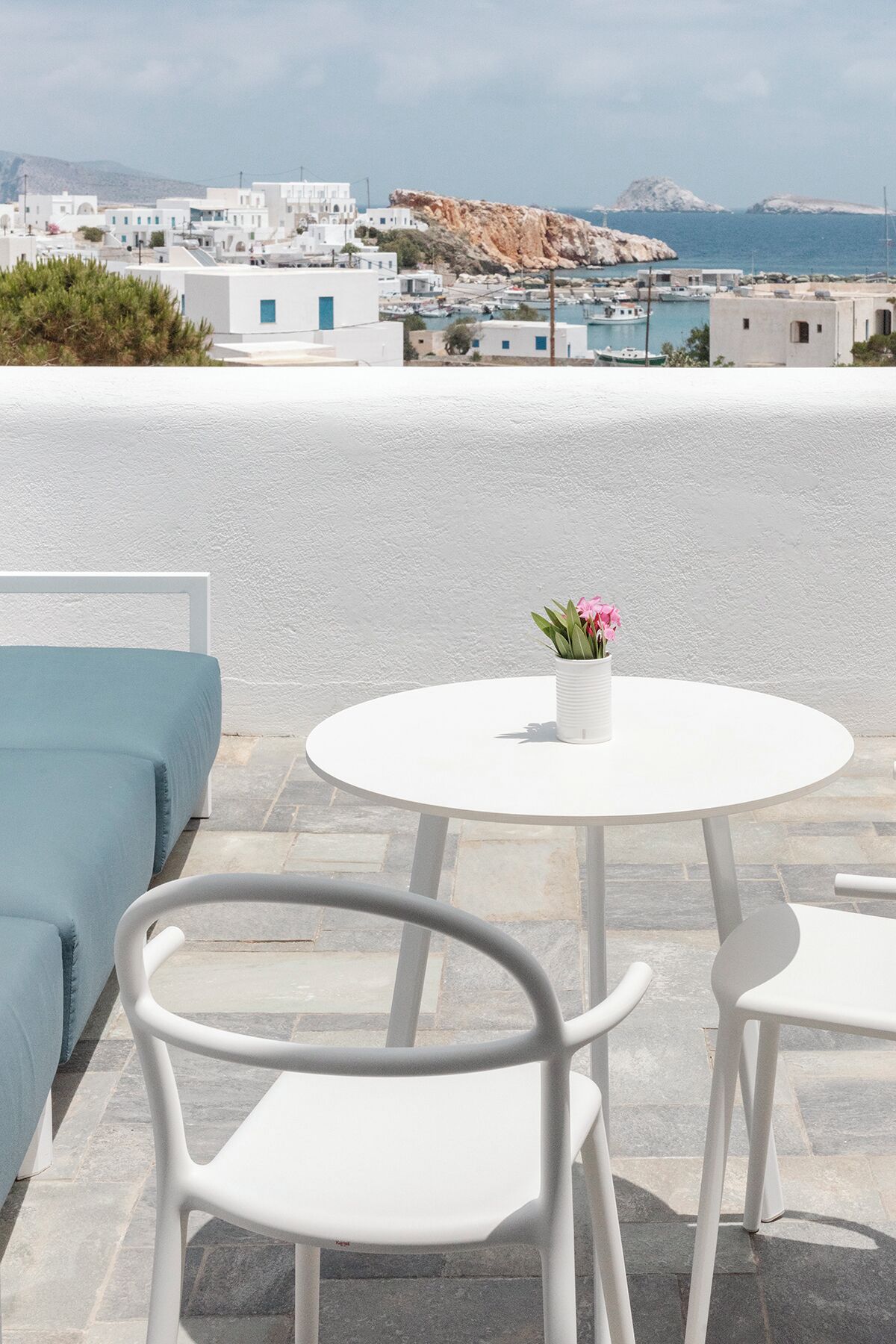 A white table with a vase of flowers, white chairs, and a blue sofa on a terrace with a view of a coastal town and buildings.