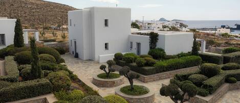 Une cour intérieure agrémentée de haies aux formes géométriques, de bâtiments blancs et offrant une vue sur la mer.