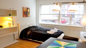 A bedroom with a bed, bedside table, lamp, window with blinds, and a view of the street. - Studio Rådmandsgade (Copenhagen)