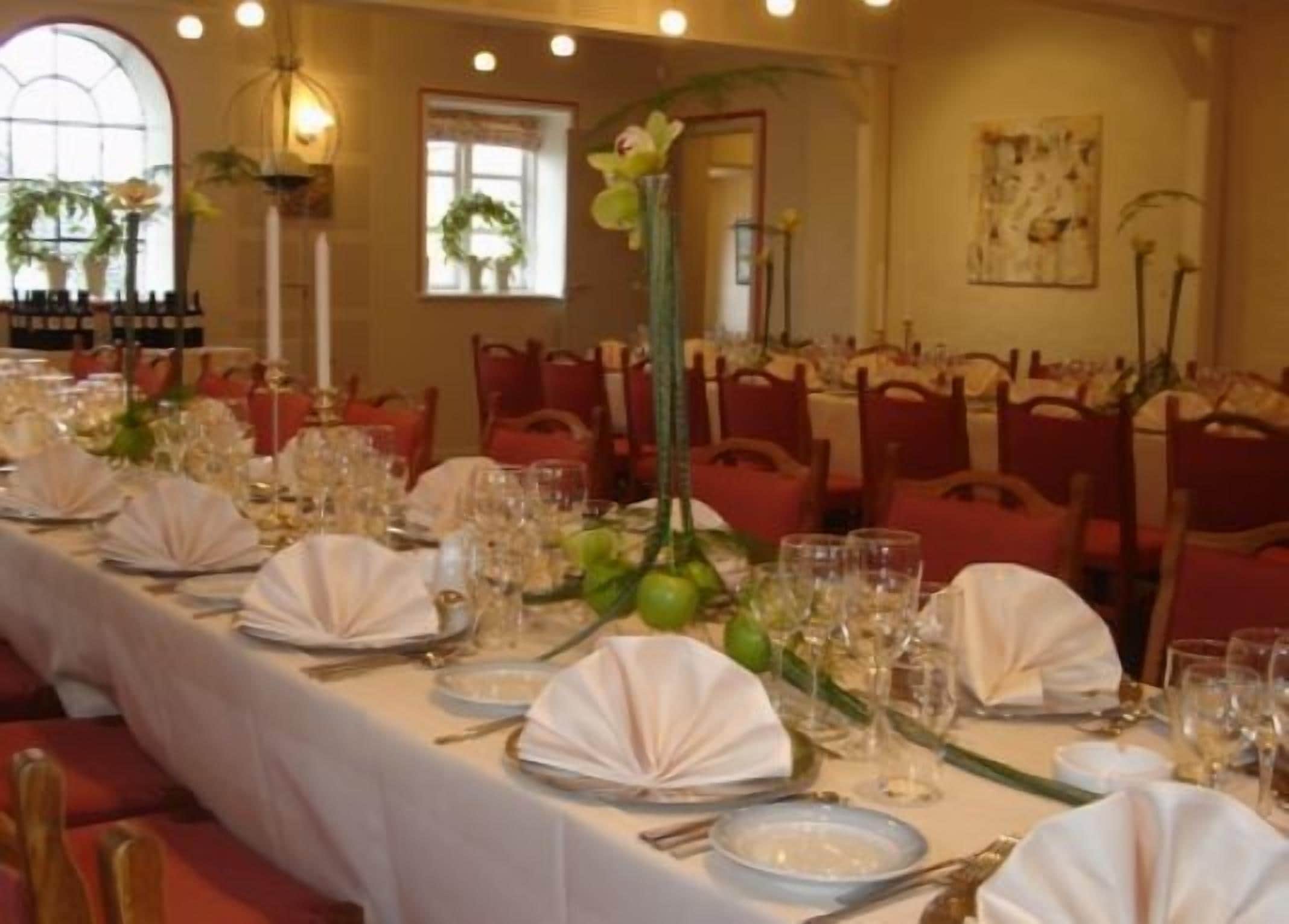 Un comedor con largas mesas dispuestas para un evento formal, con manteles blancos, cristalería y sillas rojas.
