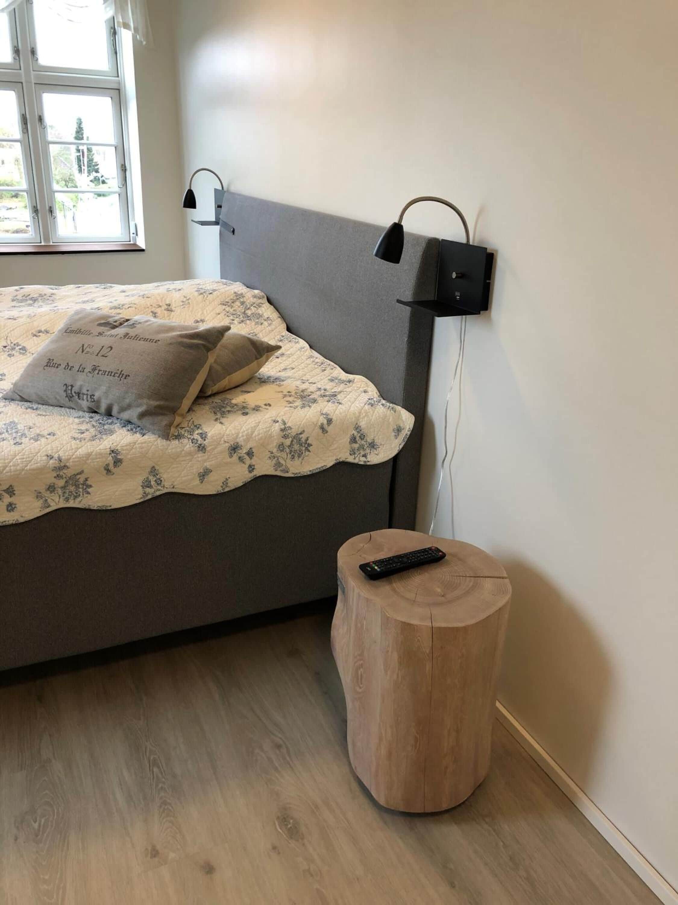 Et soveværelse med en seng, en sengetavlelampe, en lille træskammel og en fjernbetjening på gulvet.