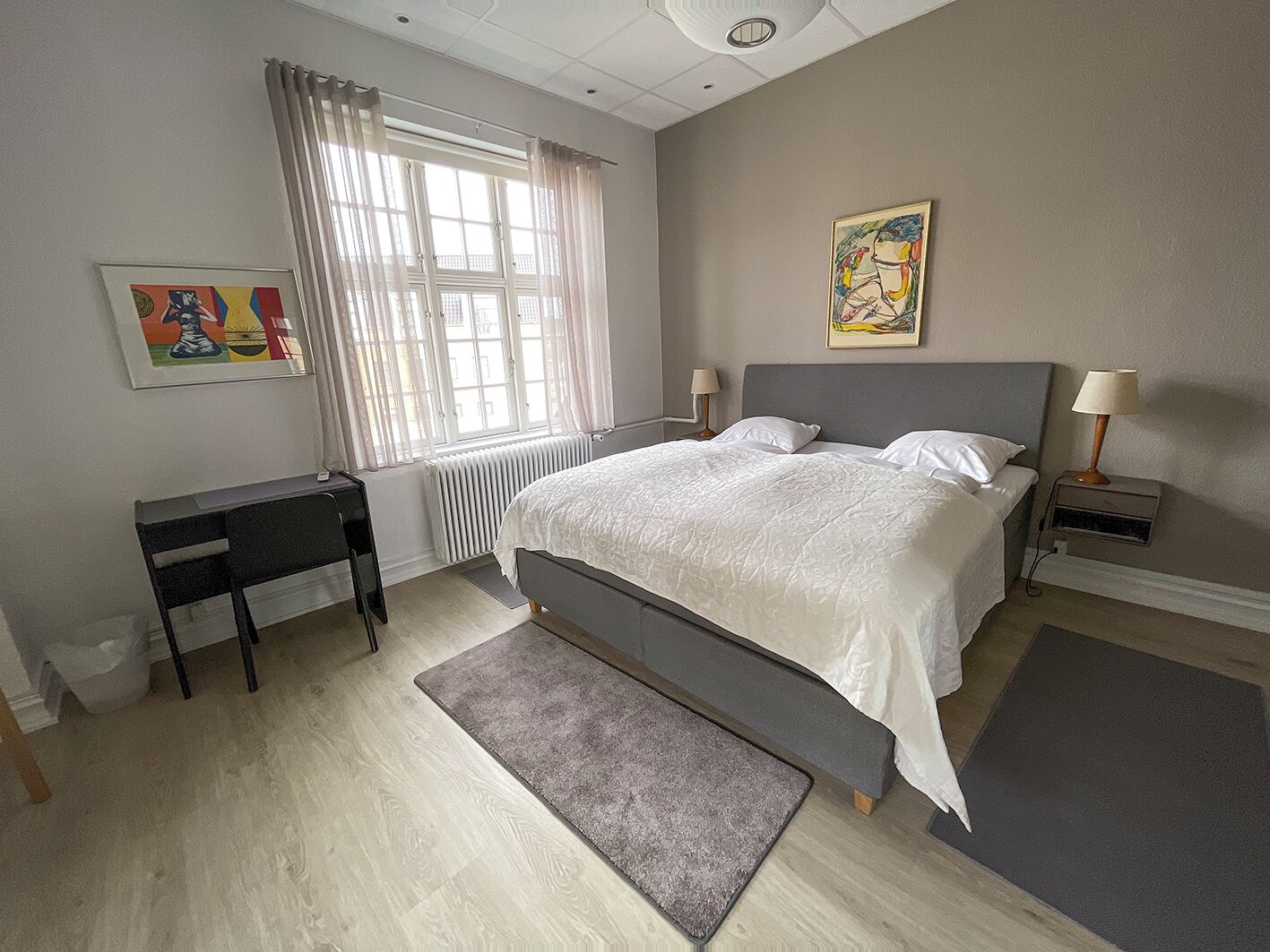 A bedroom with a bed, bedside tables, a desk, a radiator, a window with curtains, and artwork on the walls.