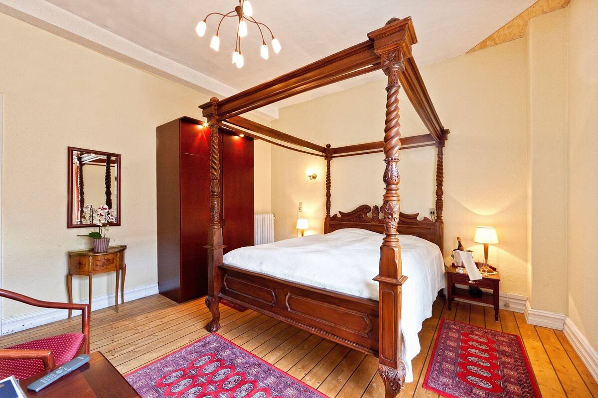 A four-poster bed with a wooden frame, a red and white patterned rug, a wooden chair, and a small table with a lamp.