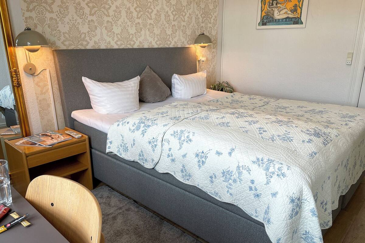 A neatly made bed with a floral-patterned bedspread, two bedside lamps, a small bedside table with a magazine, and a chair.