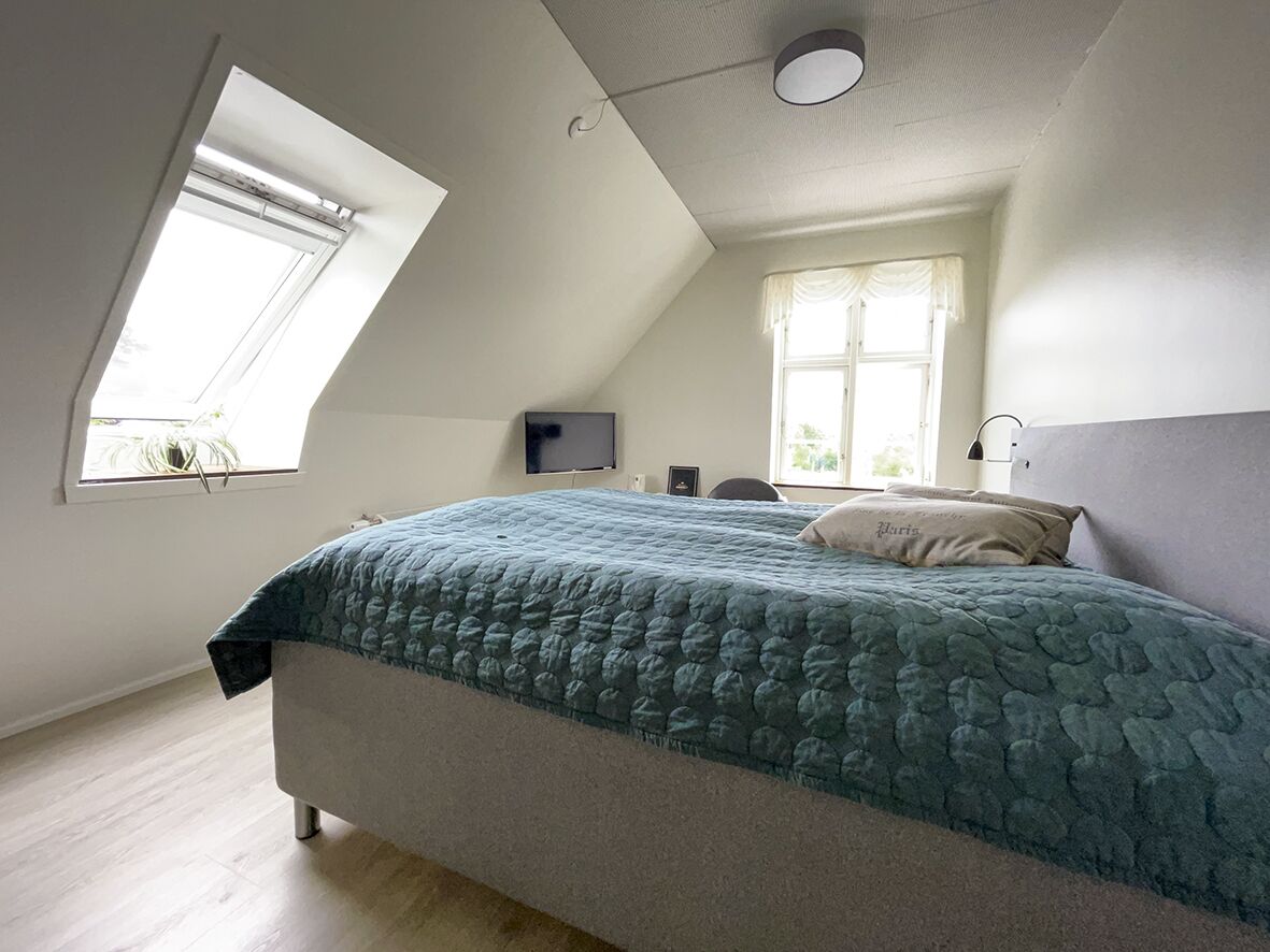 A bedroom with a sloped ceiling, a skylight, a bed with a blue quilt, a bedside table, and a window with curtains.