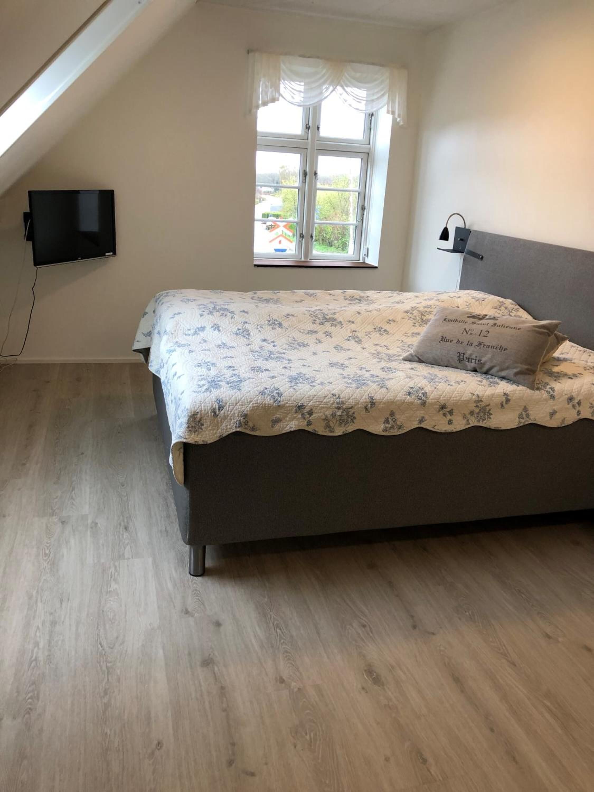 A bedroom with a bed, a window, a flat-screen TV, and a bedside lamp.