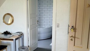 A room with a desk, chair, and a trash bin. There is a door leading to a bathroom with a white tiled wall and a bathtub. - Højslev Kro (Hojslev)