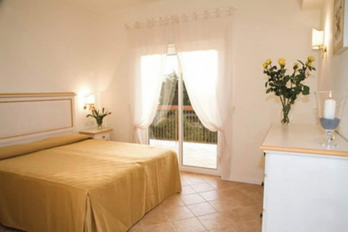 A hotel room with a bed, a nightstand with a lamp, a vase with flowers, and a view of the outdoors through a window.