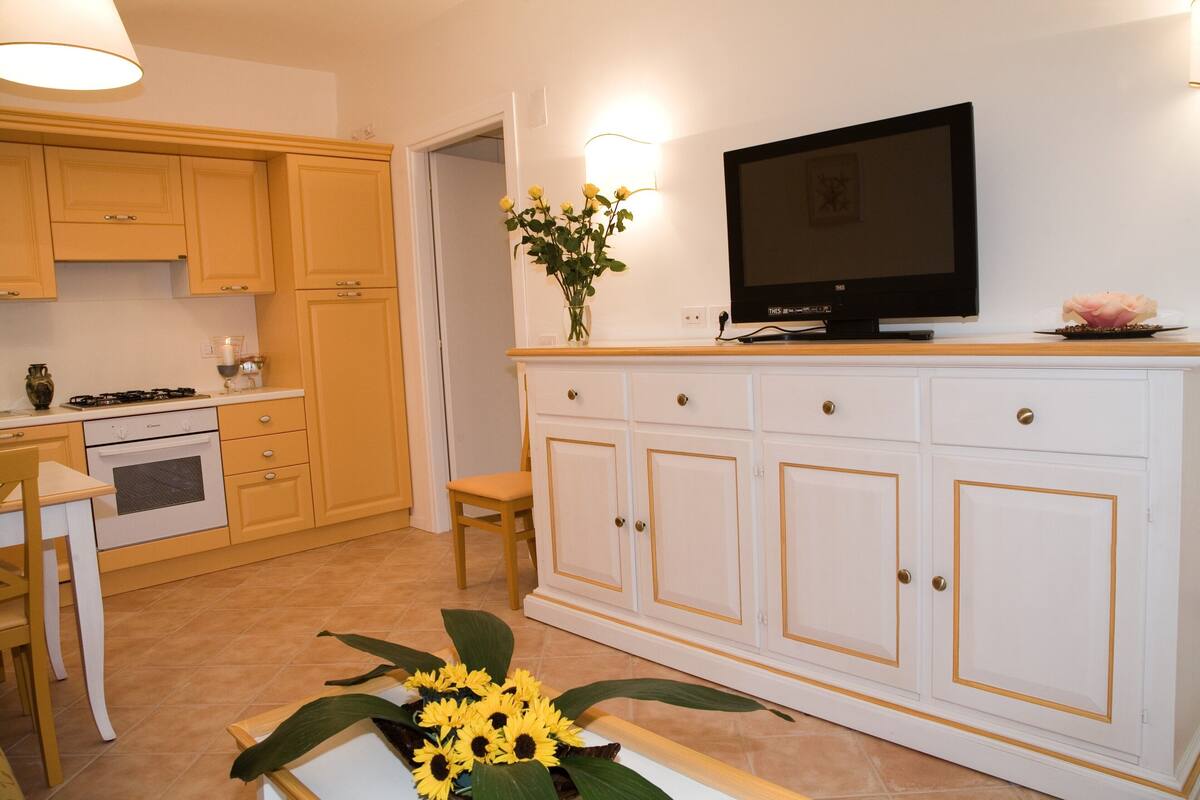 A kitchen with yellow cabinets, a white countertop, a stove, a sink, and a television on a wooden shelf.