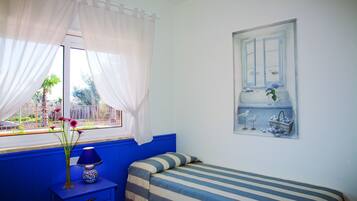 Una habitación con cama, mesita de noche, ventana con cortinas y estante en la pared con objetos decorativos.