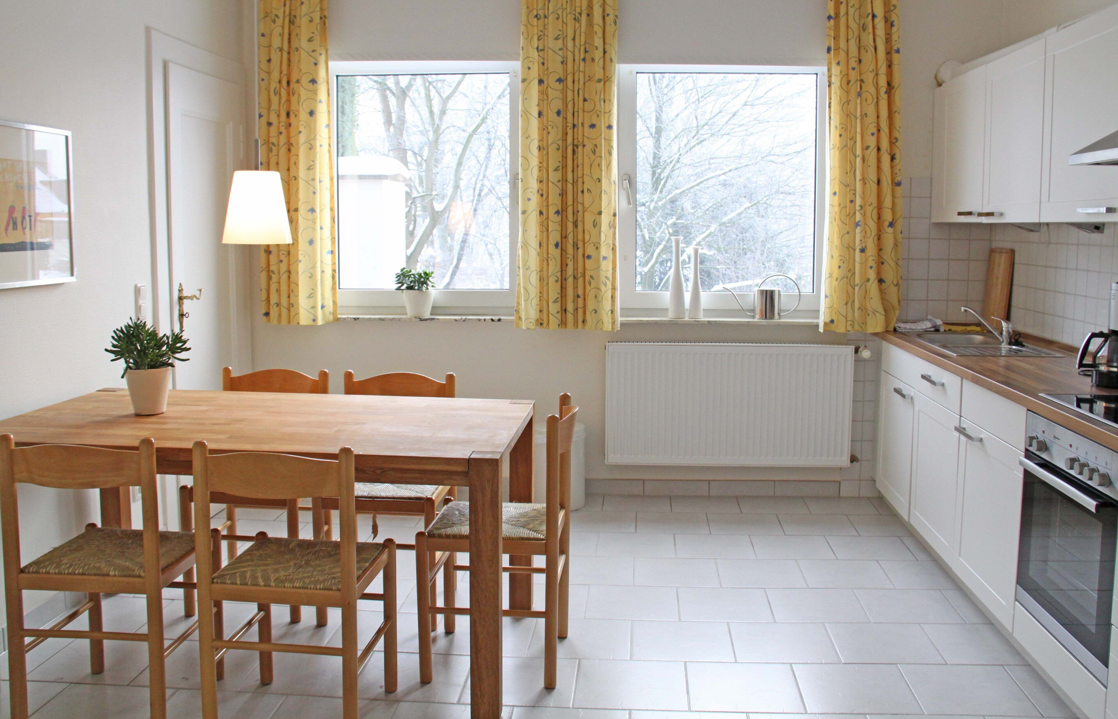 Eine Küche mit Esstisch, Stühlen, Heizkörper, Fenster mit Vorhängen und Spüle.