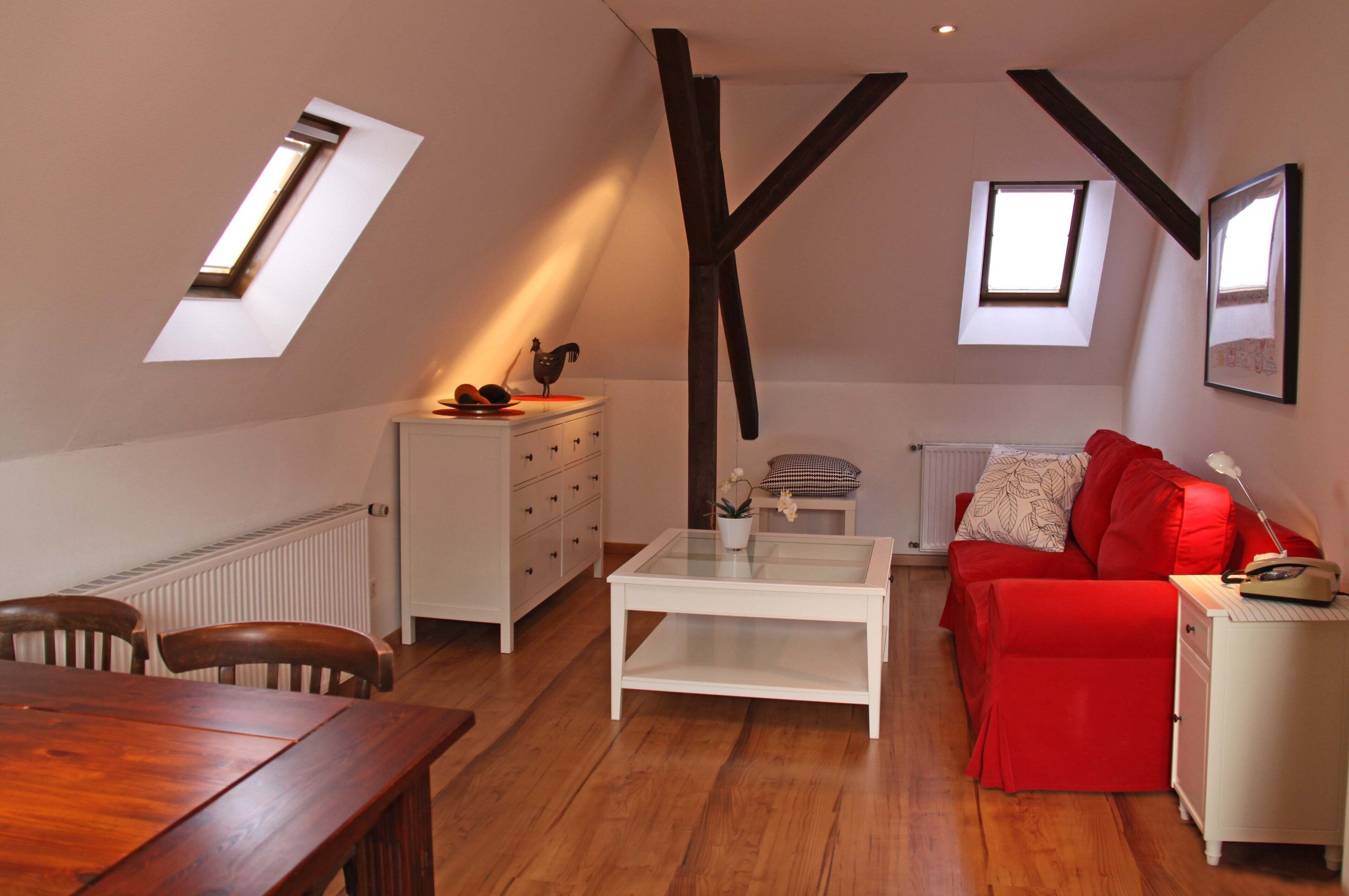 A loft-style room with a dining table, a red sofa, a white dresser, and a glass-top coffee table. There are two skylights and a radiator on the wall.