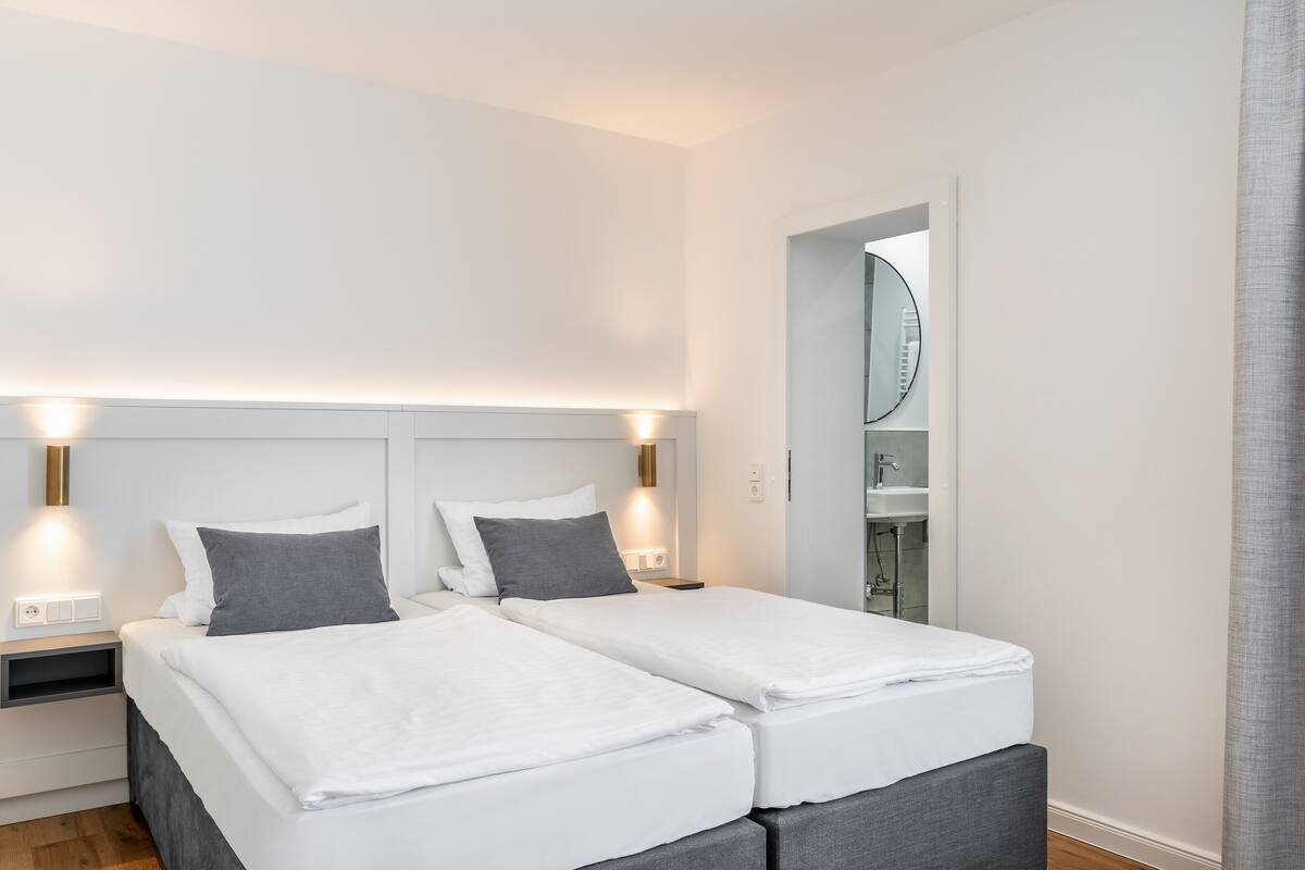 A modern hotel room with two single beds, a nightstand with a lamp, a mirror, and a bathroom visible through an open door.