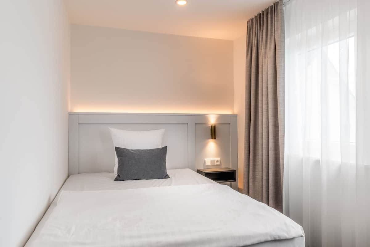 A neatly made bed with a white and gray pillow, a bedside table with a lamp, and sheer curtains on a window.