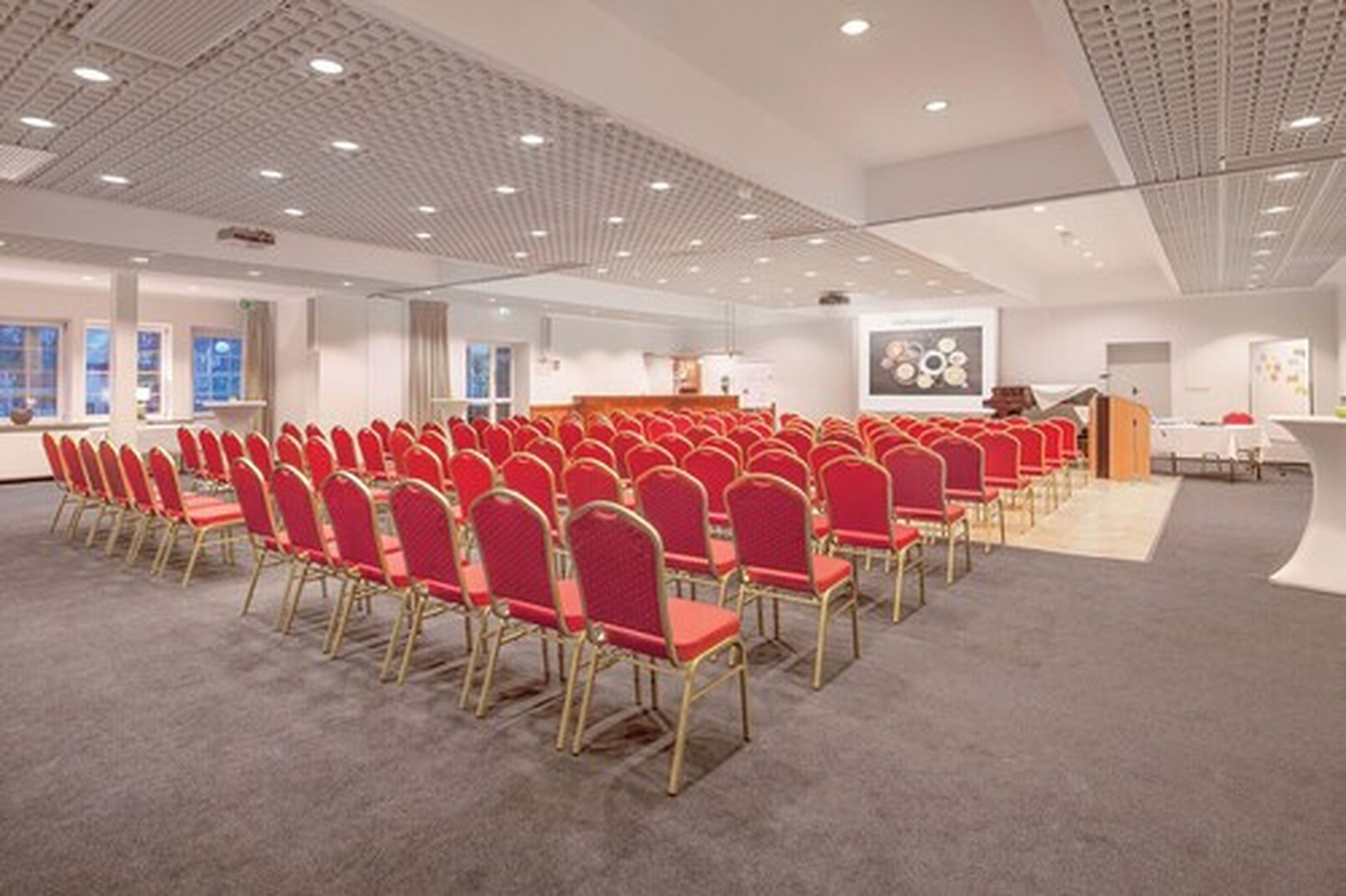 Un sal ón de conferencias con filas de sillas rojas, un atril y una pantalla de presentación.