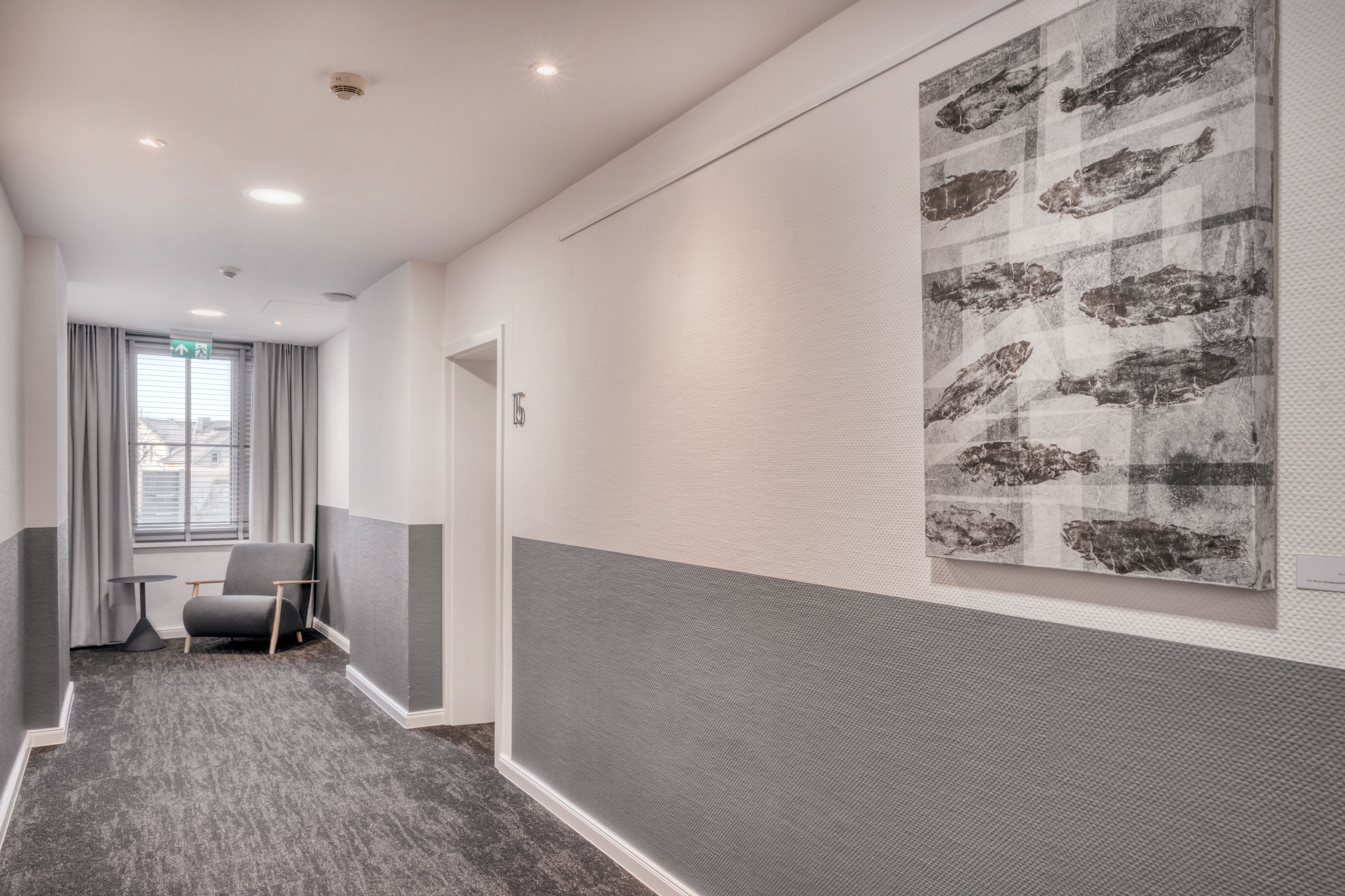 A hallway with a grey carpet, a grey armchair, a small table, and a large abstract painting on the wall.