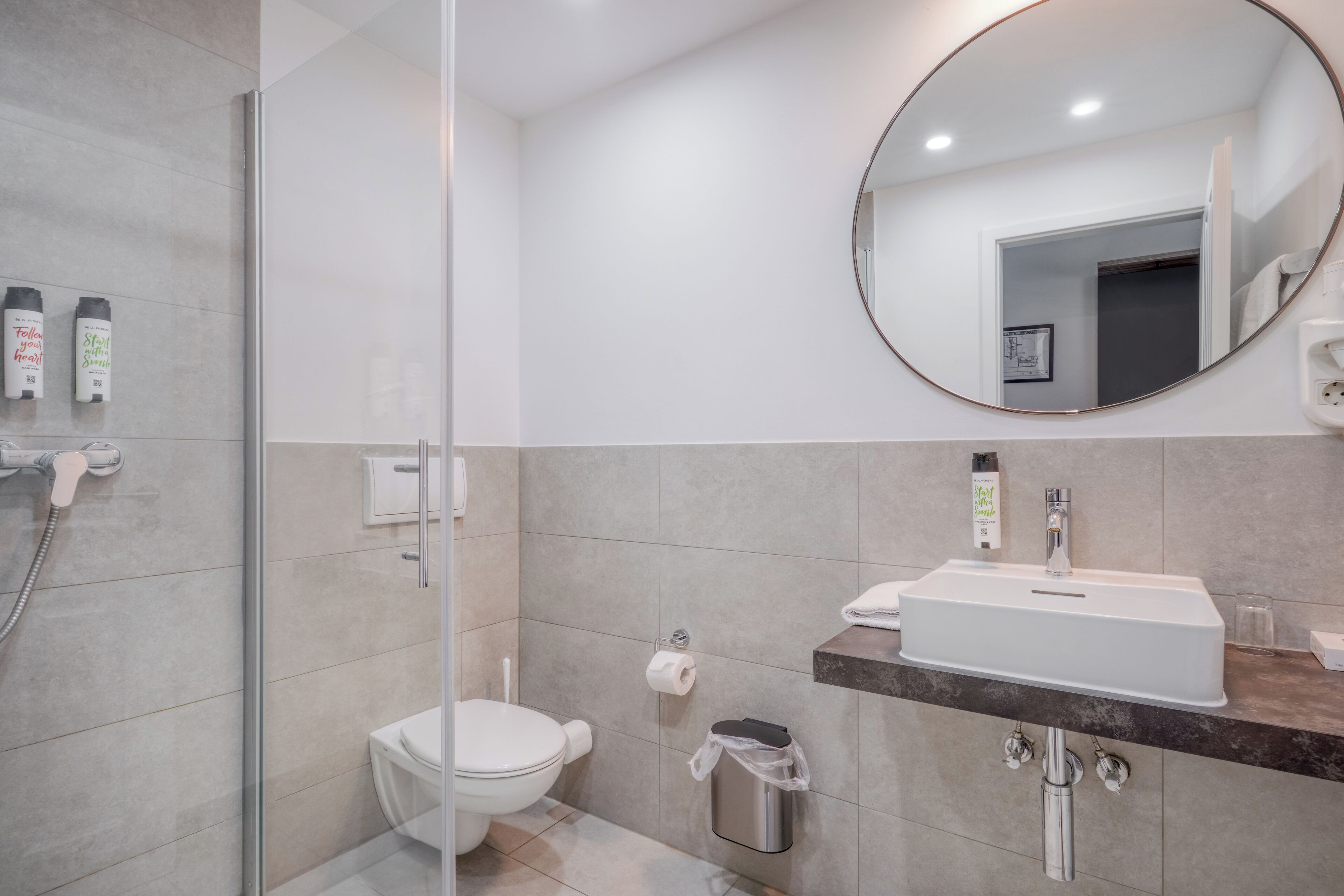 Un bany modern amb una cabina de dutxa de vidre, un vàter blanc, un lavabo amb taulell de marbre i un mirall rodó.